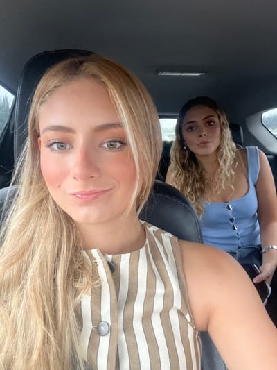 This image depicts two young adult females inside the passenger cabin of a vehicle, located in Camargo, Spain. The female in the immediate foreground, positioned in what appears to be the front passenger seat, faces the camera directly with a slight smile. She has long, light blonde hair parted centrally and light-colored eyes, wearing a sleeveless garment with vertical stripes of beige and off-white featuring a buttoned placket. The second female is situated in the rear passenger seat behind the first, her head slightly turned to her right, not directly engaging the camera. She has wavy, light-toned hair and is wearing a light blue sleeveless top with a buttoned front and a silver-toned wristwatch on her left wrist. The vehicle's interior consists of dark-colored seating surfaces. Through the windows, a blurred outdoor environment with green foliage is visible.