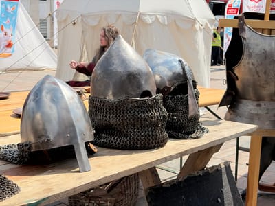 In Moskovskiy, Russia, at an outdoor event, a table displays medieval armor. Three conical metal helmets, two with attached chainmail coifs, rest on the table. To the right, a metal chest plate stands upright. A woman with long, wavy hair sits behind the table in front of a white tent, engaged in an activity with her hands. Banners with text and images are visible in the background, along with people in the distance and another tent. The scene is set on a paved ground surface.