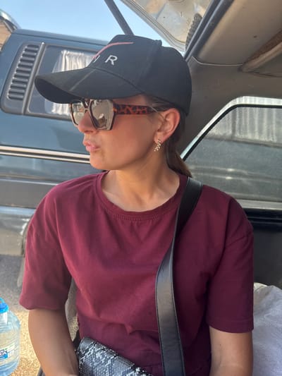In Maladzyechna, Belarus, a woman wearing a black baseball cap and oversized, leopard-print sunglasses is seated inside a vehicle. The cap has white lettering, possibly "YR". She is also wearing a maroon t-shirt and a small, delicate earring. A black strap crosses her shoulder, and a patterned clutch is visible in her lap. To her left, a large plastic bottle of water is on the ground. The interior of the vehicle, with its visible upholstery and window shade, suggests a travel or outdoor setting.