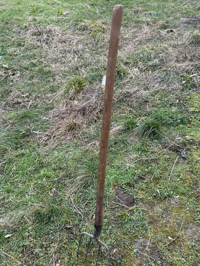 A weathered pitchfork with a long, light brown wooden handle and four dark metal tines is vertically embedded in the ground at an unspecified outdoor location within the city of Radomirtsi, Bulgaria. The ground consists of a mixed covering of vibrant green grass, extensive patches of dry, light brown grass, and visible areas of soil. Small twigs and dried plant matter are scattered across the surface. In the mid-ground, slightly to the right of the pitchfork's handle, a partially visible, translucent cylindrical object with an orange cap lies horizontally on the ground amidst the vegetation. No individuals are present, and no actions or interactions are depicted.