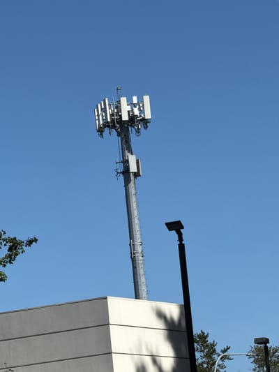 A tall, multi-faceted cell tower, a common urban structure, is captured against a clear blue sky in this image. The tower, composed of metallic segments, is equipped with an array of antennas, suggesting its function in wireless communication. In the foreground, the upper portion of a light-colored building is visible, its clean lines and horizontal bands indicating a modern architectural style. To the right of the cell tower, a black, utilitarian light pole stands, its design stark and functional. A few scattered trees with dark green foliage provide a natural contrast to the man-made structures. The scene is devoid of human presence, focusing solely on the infrastructure. The lighting suggests a bright, daytime setting, casting subtle shadows. The overall impression is one of an urban environment, characterized by a blend of technology and modern architecture.
