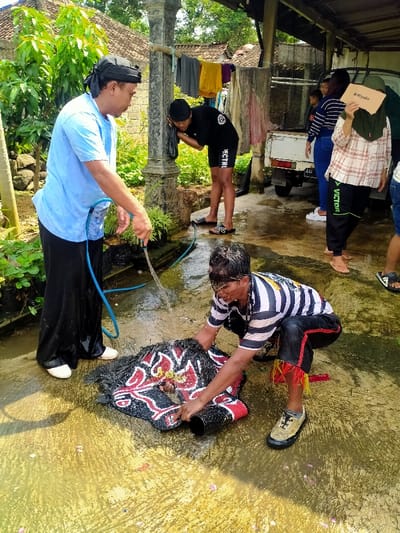 A man in a blue shirt and black pants holds a hose and sprays water onto a red, black, and white patterned rug on the ground. Another man in a black and white striped shirt is rinsing the rug. There are four women standing behind the man, two of whom are holding onto the rug and a wooden door behind them. There are some flowers on the ground around the rug. The scene is set in the courtyard of a home with a concrete floor and a partially covered walkway. The hose is connected to a faucet that is mounted on a large gray post. The location is a residential area in Kecamatan Kedawung, Indonesia, with coordinates -7.52 latitude and 111.028 longitude.