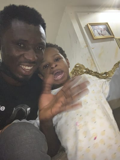 This image depicts an adult male and a young child in a close-up indoor setting, consistent with a residence in Kurudu, Nigeria. The adult male, positioned on the left, has dark skin, short dark hair, and is smiling with visible teeth. He wears a dark t-shirt with a partial white graphic and dark grey trousers; a dark strap is visible near his neck. The child, to the right of the adult, has dark hair, lighter skin, and an open mouth revealing teeth. The child wears a light-colored, long-sleeved top featuring a yellow pattern and purple stains, and one hand is raised and blurred, indicating motion. The background features a light-colored wall with visible blemishes, decorative molding, a gold-framed picture containing three figures, and a segment of ornate, gold-colored furniture on the right. Specific location details within Kurudu are not visually discernible.