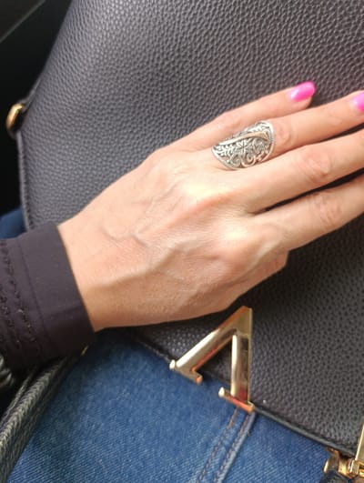 The image shows a close-up of a woman's hand resting on a black textured bag and blue denim. The woman is wearing a silver ring with an intricate floral design on her right index finger. Her fingernails are painted a bright pink. The bag has a prominent gold metal accent on the front. The background is out of focus but appears to be indoors. There is no visible indication of the city of Penza, Russia, nor is there any context provided to infer its location.