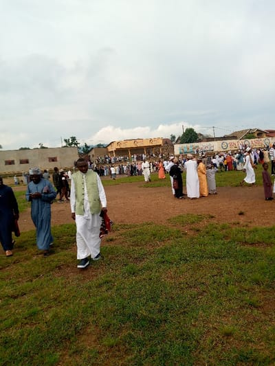 The image shows a large gathering of people in an open field in Beni, Democratic Republic of Congo.  The field is covered in green grass and has a dirt track running through the middle.  There are several buildings in the background, including a large, yellow building with a sign. The sign is in Arabic and reads, "BNI ASBL" .  The people in the image are mostly men, and many of them are wearing traditional white robes.  They are standing around in groups, talking and laughing.  It appears to be a celebration of some kind, with food and drinks being served.  The people appear to be in a celebratory mood, with the air filled with excitement and joy. The sky is cloudy, and the weather appears to be humid and warm. The image is taken from a low angle.