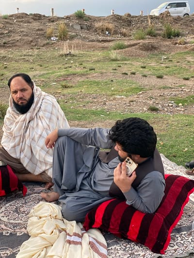 Two men are observed seated outdoors on a patterned rug in Arghandab, Afghanistan. The man on the left, with a full dark beard, is wrapped in a beige and brown plaid shawl over light brown attire, looking directly forward. The man on the right, with dark curly hair and a short beard, wears a grey long-sleeved garment and a dark vest, seated on a red and black striped cushion. He holds a light-colored smartphone to his right ear, seemingly engaged in a call. His left foot is bare. On the rug in front of him is a pile of cream-colored fabric with brown patterned stripes. The background consists of a dry, rocky, and sparsely vegetated terrain featuring a dirt mound topped with three white and red posts. A light-colored minivan is partially visible on the far right of the mound. The sky is overcast.