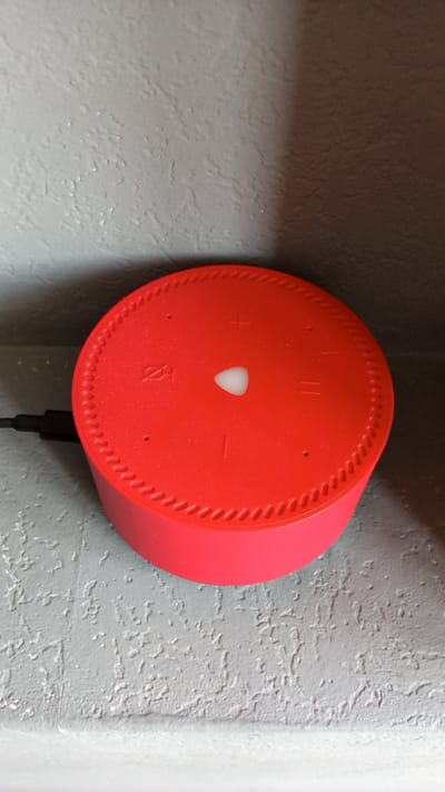 A red Amazon Echo Dot device sits on a shelf. The device has buttons for volume up, volume down, microphone mute, and an action button. A power cable is plugged into the back of the device. The shelf is painted a light gray color and has a textured surface. The background is dark. The image was taken in Aderbiyevka, Russia.