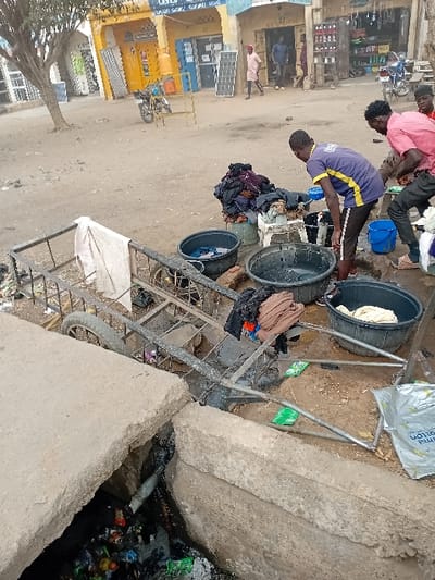 The image depicts an unpaved street scene in Sharada, Nigeria. In the immediate foreground, an open concrete drainage ditch runs horizontally, filled with various debris including plastic bottles and wrappers. A metal pushcart with two wheels is positioned over the ditch, with a white cloth hanging from its side. On the dusty ground, three large black plastic tubs are arranged, some containing water and clothes.
Two adult males are the primary subjects in the midground, engaged in laundry activity. One male, wearing a blue and yellow short-sleeved t-shirt and dark pants, is bent over a tub, holding a small blue basin, appearing to be washing or rinsing garments. To his right, a second male, dressed in a red short-sleeved t-shirt and dark pants, is crouched near another tub. A third male in a red shirt is partially visible behind the second individual. A pile of folded or bundled clothes rests on a white stool.
The background features a row of single-story commercial buildings with facades in shades of yellow and light blue. Various retail items, including solar panels and a gas cylinder, are visible outside the establishments. Two motorcycles are parked on the unpaved street. Several other individuals are observed standing or walking further down the street near the shops. Trees with sparse leaves are present on the left side of the frame. The lighting suggests daylight conditions.