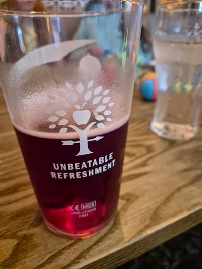 The image depicts a pint glass filled with a red beverage, likely a dark beer or cider, sitting on a wooden table. The glass has a frosted condensation on the exterior and a white tree-shaped logo with the words "UNBEATABLE REFRESHMENT" printed beside it. A CE marking and "0866 Utopia PINT" are visible near the base of the glass. In the background, another glass with a clear liquid, presumably water, is partially visible. A multi-colored spherical object is also vaguely seen in the background, sitting on the wooden table. Given the type of drink and the typical pub setting, it's likely located in a pub setting, the details suggest it might be in Breckland District, United Kingdom.