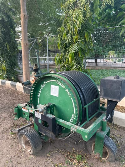 This image depicts a green, industrial-sized water reel with a large coil of black hose. The reel is mounted on a green metal trailer with four wheels, two of which are visible in the foreground and appear to be standard tires, while the rear tires are agricultural or turf-style with deep treads. A black rectangular tank is attached to the right side of the trailer. The reel has "Water-Reel" printed in white script on its side, along with a label containing technical information. There is also a "WARNING" label visible.
The trailer is positioned on a dirt and gravel surface, partially covered with fallen leaves. A concrete curb with black and white stripes runs along the left side of the scene. Behind the trailer, a chain-link fence encloses an area with green vegetation, possibly grass or low-lying plants. Several trees with dense green foliage are present in the background, including a prominent tree trunk to the left of the reel. The overall setting suggests an outdoor industrial or agricultural area. The specific location in the city of Batagarawa, Nigeria, is not visually identifiable from the image content alone, as there are no landmarks or distinguishing features that can be definitively linked to that specific geographic location.