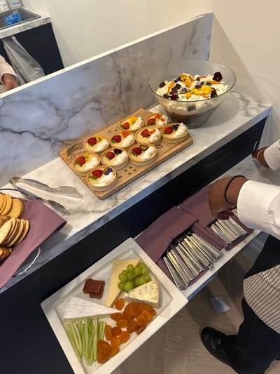 The scene depicts an indoor catering or buffet setup in Epsom and Ewell, United Kingdom. A marble-patterned countertop serves as a display surface, featuring a large glass trifle bowl filled with layers of dessert, topped with whipped cream, dark crumbs, and an assortment of yellow, purple, and reddish edible flowers. Adjacent to the trifle, a wooden board holds nine small tarts, each topped with white cream, a sliced strawberry, and small edible flowers. To the left, a set of silver tongs rests next to stacks of round crackers placed on a purple fabric napkin. Below this counter, a dark countertop provides a second display level. Here, a rectangular white platter showcases a variety of food items: a wedge of brie cheese, a slice of a light yellow cheese, a wedge of blue cheese, a cluster of green grapes, several dried apricot halves, and cut celery sticks. Another rectangular white tray contains multiple sets of silver cutlery (forks and spoons visible), each neatly rolled within a purple fabric napkin.

Two individuals are partially visible. On the left, a person wearing a light-colored long-sleeved shirt is seen reaching into an area behind the marble counter, with a clear plastic bag visible in the background. On the right, a person in a white long-sleeved shirt, black trousers, and a brown checkered garment (possibly an apron or lower part of a uniform) is standing next to the lower counter. Their right hand, adorned with a black hair tie on the wrist and a gold bracelet, is positioned above the cutlery tray. Their black dress shoe is visible on the light wooden floor beneath the counter.