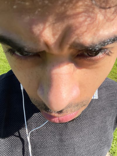 A high-angle, close-up photograph captures an individual outdoors in Montigny-le-Bretonneux, France. The subject, identified as an adult male with a medium-dark complexion, dark hair, and stubble/mustache, occupies the majority of the frame. His forehead is furrowed, and his dark eyes are directed downwards. He is dressed in a dark grey, finely knitted sweater layered over a light-colored, collared shirt with a subtle plaid or striped pattern. White wired earphones are visible, with a wire descending along his left side. The background is a blurred expanse of green grass, indicative of an outdoor setting. The scene is illuminated by strong, direct sunlight, casting distinct shadows on the subject's face and clothing.