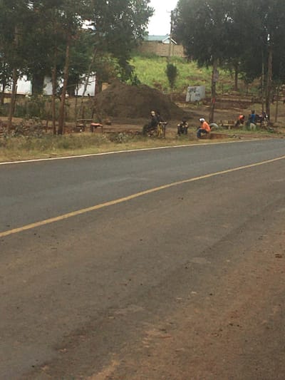 Along a two-lane road with a yellow center line, several people are engaged in roadside work. One person in an orange shirt is bending over, while others are near a dirt mound. Bicycles lean against the mound. A grassy area lies beyond the workers, with trees lining the road. A building is partially visible in the distance. The road surface transitions from a dark, smooth asphalt to a rough, brown material closer to the viewer.