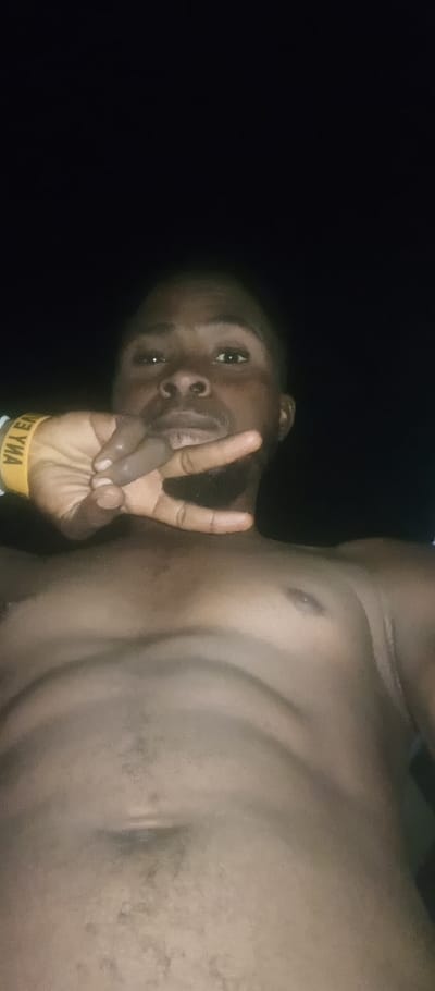 A shirtless, dark-skinned man poses in a selfie. His face is clearly visible, and he is holding up a hand with fingers forming a peace sign near his face. He has short hair and a slight beard. The lighting is poor, with a dark background. He wears a yellow wristband. The camera angle is low, capturing his chest and torso. The man appears to be posing confidently.