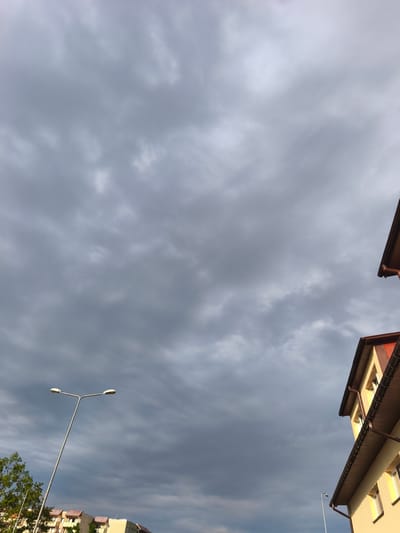This media file depicts an overcast sky with visible clouds. In the foreground, a streetlamp with two lights stands prominently, its metal pole angled upwards. To the right, the eaves of two buildings, characterized by their brown trim and reddish-brown roofs, are visible. One building is a light yellow color with visible windows. In the background, more buildings with similar architectural features can be seen, along with the top of a green tree on the left side. The overall scene suggests an outdoor setting in an urban or suburban environment, likely during the daytime given the lighting. The cloudy sky and the muted colors hint at potentially gloomy or preparing-for-rain weather. There is no visible text in the image. The setting is identified as Szczecin, Poland.