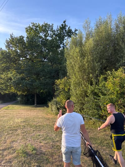 Two young men are on a grassy area with trees in the background. One man in a white t-shirt and denim shorts is facing away from the camera, with his left arm raised. The other man, wearing a black tank top and athletic shorts with yellow stripes, is facing left and appears to be holding a fishing net and a fishing rod. The environment suggests an outdoor recreational area. The image's content, specifically the presence of a fishing net, suggests the activity might be fishing. This scene is consistent with various recreational spots along the Rhine river in Ludwigshafen am Rhein, Germany, known for its parks and waterfront areas suitable for outdoor activities.