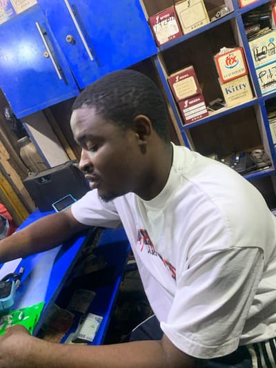 An indoor scene, likely a shop or business in Garaku, Nigeria, features a man positioned centrally, looking downwards to his right. The image is taken at an oblique angle, making the man and the environment appear tilted. He is a dark-skinned individual with short, dark hair and a neatly trimmed beard or goatee, wearing a white short-sleeved t-shirt with some red and black graphic designs on the front.
Behind the man, a wall of blue-painted wooden shelves is visible, filled with numerous boxes and items, suggesting an inventory of goods. Some boxes have discernible labels, including "KITTY," "LCD CAMBO LCD," and "TOUCH," along with a red and white circular logo displaying "LCB Touch." Below these shelves, there are blue cabinet-like structures with drawers or compartments.
To the man's lower right, on a counter or shelf, stacks of Nigerian currency are clearly visible, with "1000" printed on the top notes, indicating 1000-naira denominations. Beside the money, a black electronic device, possibly a smartphone, rests. Further to the right, a light blue container or box with a handle is partially visible. The lighting is artificial, illuminating the foreground and parts of the shelves, while some areas are darker. The overall context suggests a retail or service environment, possibly dealing with electronics or phone parts, where the man is engaged in a transaction or task.