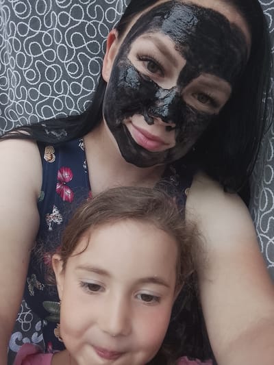 This candid medium-shot captures a woman and a young girl in a relaxed, intimate setting, identified as Kuyeda, Russia. The woman, positioned in the upper part of the frame, has long dark hair and is wearing a dark blue dress adorned with a vibrant butterfly pattern in shades of pink, brown, and white. Her face is mostly covered with a shiny, thick black charcoal mask, leaving her light brown eyes with prominent eyelashes, nose, and mouth area partially visible. She has a soft, slight smile on her lips.
Below and slightly in front of the woman is a young girl, whose light brown, wavy hair frames her partially visible face. She appears to be wearing a pink top. The girl is looking downwards and to the side, exhibiting a gentle, perhaps thoughtful or slightly playful expression, with her lips slightly parted.
The background consists of a grey fabric patterned with a repeating abstract design of white swirling or looping lines, possibly indicating a curtain, blanket, or furniture upholstery. The natural lighting suggests it is daytime. The scene conveys a warm, casual moment, likely a self-care routine like a face mask, with the child nearby. No visible text is present in the image.