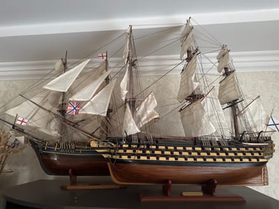 The image shows two detailed model ships displayed on a shelf. The ship in the foreground is larger and has a black hull with yellow detailing, and numerous cannons visible along its side. The ship behind it is smaller and has a dark blue hull with a yellow stripe. Both ships have white sails, some of which are partially furled, and rigging. Several flags are visible on the ships, including what appear to be British flags. The models are mounted on wooden stands. The background consists of a wall with floral wallpaper and a white ceiling with molding. There is no indication of the city of Давыдовка, Ukraine in the image; this is a domestic setting.