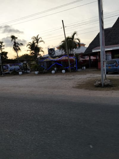 The image depicts a roadside scene at dusk or dawn in Gombato Bongwe ward, Kenya. In the foreground, a dark asphalt road occupies the lower portion of the frame. Beyond the road, an unpaved, light brown dirt area serves as a shoulder or parking space.

In the midground, a commercial establishment is visible. On the left, a large banner, partially obscured, displays the word "KARAOKE." Adjacent to this, a building identified by a circular sign as "The Club Restaurant" with "ACCOMMODATION" beneath it, features a white roof and is decorated with prominent blue string lights strung in wavy patterns along its facade. Several white spherical planters containing green plants are arranged in front of this building. Palm trees are scattered around and behind the structure.

To the right of "The Club Restaurant," another building with a distinctive dark thatched roof is present. A red awning extends from this building, and a partial sign on its exterior appears to read "PIZZA." A blue compact car is parked facing left in front of this thatched-roof structure. A tall, light-colored utility pole stands vertically on the far right, with overhead power lines crossing the sky.

The background shows a sky transitioning from a pale blue at the top to a warm yellow-orange hue near the horizon, indicating either sunrise or sunset. Sparse clouds are present. No individuals are visible in the scene.