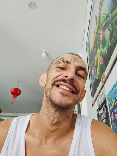A man with a beard and visible tattoos is smiling broadly and looking at the camera. He is wearing a white tank top. A framed picture depicting lotus flowers and a blue sky is on a white surface in the foreground. There are advertisements and magazines, one with the word "MÁYLO" visible, next to the framed picture. A red lantern is visible hanging from the ceiling in the background. The image may have been taken in Cam Ranh Ward, Vietnam, based on the language of the advertisement. The room appears to be indoors, with white walls and a light fixture.