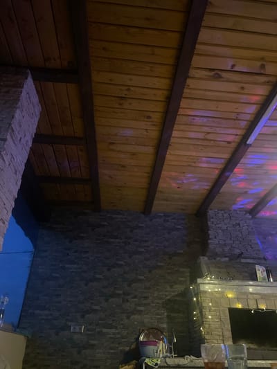 This image captures an indoor or covered outdoor entertainment area, located in Petrivka, Ukraine.
The scene is illuminated by dim artificial lighting, creating a warm yet shadowy ambiance. The ceiling is constructed from horizontal wooden planks, supported by evenly spaced dark brown wooden beams. Dynamic, multi-colored light reflections (red, blue, and purple) are cast across the right half of the wooden ceiling, indicating the presence of a rotating party light source, such as a disco ball, located out of frame to the right. A single white linear light fixture is partially visible along the rightmost beam.
The walls are clad in dark grey, textured stacked stone veneer. On the far left, a stone pillar or structural element extends upwards. A subtle blue light emanates from an area in the bottom left corner, possibly from a window or another light source. Towards the lower left of the back wall, a standard electrical outlet with two receptacles is visible.
On the right side of the image, a substantial built-in structure, likely a fireplace hearth or an outdoor kitchen unit, is constructed from the same stacked stone. This structure is decorated with warm white string lights draped across its front and along its lighter-colored top surface. On this top surface, several indistinct objects, including what appears to be a framed picture and a dark bottle, are visible. Below the top surface, a dark rectangular opening, consistent with a firebox or grill cavity, is present.
In the immediate foreground on the left, a countertop surface holds various items, including a metallic serving tray, a purple plastic bowl, a glass, and a faucet, suggesting a sink area or bar preparation station. Directly in front, two prominent drinking glasses are positioned on a surface. The left glass contains an amber-colored liquid, possibly an alcoholic beverage, while the right glass holds a clear liquid. No individuals are visible in the scene.