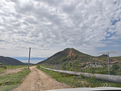 This image captures a dirt road winding through a rural landscape, leading towards a body of water framed by rolling hills. A large, weathered pipe runs parallel to the road on the right side, separated by a wire mesh fence. In the distance, the lake of Issyk-Kul is visible, with a small town nestled on its shore. The sky is overcast with scattered clouds, casting a diffused light on the scene. Power lines and a utility pole are also present along the road. The overall impression is one of a remote, natural setting with signs of industrial infrastructure.