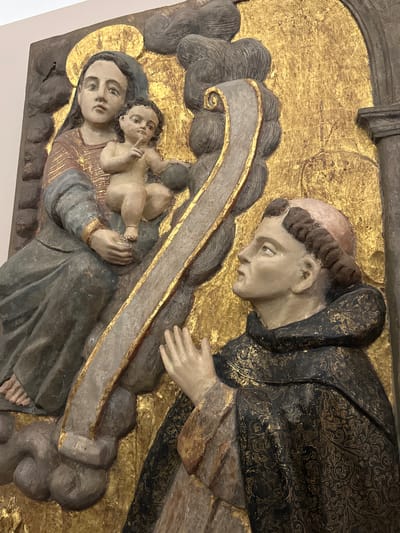 This media file displays a religious relief carving, likely part of an altarpiece or devotional artwork, from Tepotzotlán, Mexico. The carving features three central figures against a richly gilded background.
On the left, a woman, presumably the Virgin Mary, is depicted holding an infant, likely Jesus Christ. She wears a dark blue head covering and a reddish-brown tunic. A faint halo is visible behind her head. The infant, with a somewhat curious expression, raises one hand and looks upwards.
To the right, a male figure is shown, presumably a saint, perhaps a Franciscan friar given his tonsured haircut and habit. He wears a dark, almost black, habit richly adorned with intricate gold brocade patterns. He gazes reverently towards the Virgin and Child, with his hands gently reaching out or clasped in adoration.
The figures are surrounded by dark, swirling cloud-like motifs, and a light grey ribbon or scroll with gold trim winds diagonally between the main figures. The background is covered in golden leaf, which appears textured and somewhat aged, suggesting a celestial or sacred setting. The overall style points to a Baroque or Colonial era artwork, characterized by detailed carving, rich ornamentation, and religious subject matter. No visible text is present in the image.