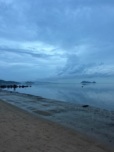 The scene presents a view of a beach in Baan Sri Thanu, Thailand. The sky is cloudy, with visible cloud formations above water with mountainous land masses in the distance. The foreground features a sandy beach area, partially wet from the receding water line, on the lower right section of the image.