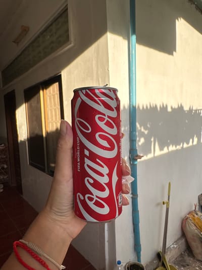 A hand holds a red and white Coca-Cola can in front of a building with white walls and a tiled floor. The can has the Coca-Cola logo and "FIFA WORLD CUP" text. Sunlight casts shadows of foliage onto the wall. A blue pipe runs vertically on the wall. In the background, a broom leans against the wall and a bag of materials is visible. The overall setting appears to be outdoors in a residential or commercial area in Siem Reap, Cambodia.