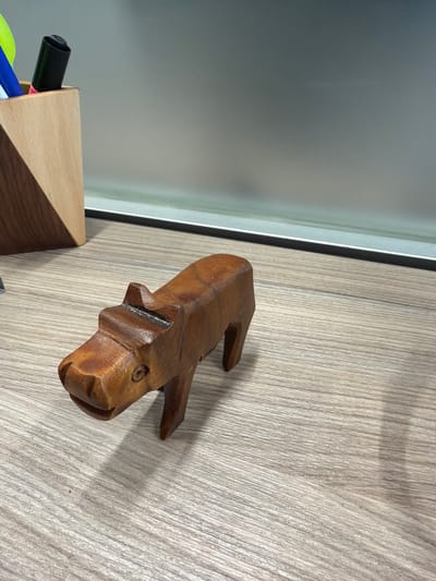The image depicts an indoor scene on a light wood-grain desk surface in Juriquilla, Mexico. A small, carved wooden hippopotamus figurine is positioned prominently in the foreground, facing towards the lower-left. The figurine is light brown with visible wood grain, exhibiting a stylized, blocky form with a distinct snout, small ears, and circular carved eyes. To the left, a light-colored wooden pen holder, constructed with a diagonal wood panel, contains a black marker with a pink end cap, a blue pen, and a partial green object. The background consists of a blurred, uniform light gray wall or panel, separated from the desk surface by a thin, light-colored horizontal line. No individuals or dynamic actions are present.