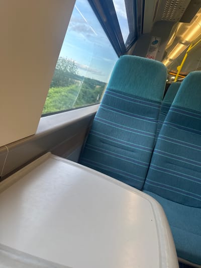 A view from inside a train carriage, looking out of a window. The seats are upholstered in a teal fabric with horizontal purple and blue stripes. A white tray table is in the foreground, angled towards the viewer. Outside the window, a green landscape with trees is visible under a cloudy blue sky. The image appears to be taken during daylight. The location of Tonbridge and Malling, United Kingdom is not discernible from the image.