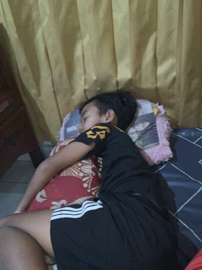 The image shows a person sleeping on a bed. The person is wearing a black shirt with yellow accents and black shorts with white stripes. They are lying on their side, with their head on a pillow. A patterned pillow and a framed photograph are visible under the person's head. The bed appears to be covered with a dark blue and white patterned sheet. The background features a wall with horizontal, textured panels. The scene suggests a casual, indoor setting, possibly a bedroom. There is no indication of the time of day or weather conditions. No visible text is present in the image. The location context is Sukasada, Indonesia.