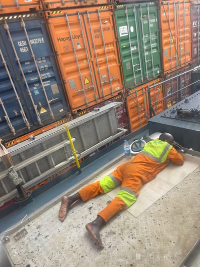 The image shows a person lying down on a metal surface, appearing to be asleep. They are wearing orange overalls with reflective yellow stripes on the legs and arms. A white hard hat is placed on the ground next to them, along with a bottle of water. The setting is a port, with stacks of large shipping containers visible in the background. The containers are colored orange, green, and blue, with various markings and labels, including "Hapag-Lloyd," "UASC," and container identification numbers. A metal ladder is also present in the scene. The lighting suggests it is daytime. The presence of the shipping containers, ladder, and the person's work attire indicate this is likely a work environment, possibly a port or terminal. Some visible text includes company names and container codes, such as "GESU 6432794 45G1", "Hapag-Lloyd", and "UASC". The overall impression is one of rest or a break during work in a busy industrial setting.
