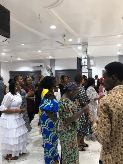 The image depicts an indoor gathering of approximately 20-30 adults, primarily women, within a venue in Gbawe, Ghana. The individuals are standing, many facing leftward from the viewer's perspective, and appear to be engaged in an activity involving movement and interaction, possibly dancing or swaying, with several women gesturing with their hands. Attire is varied, featuring traditional Ghanaian fabrics in vibrant patterns and colors, alongside more contemporary formal wear; notable garments include a white tiered lace dress on a woman in the left foreground, and a patterned brown and tan shirt on a man in the right foreground, partially obscured.

The environment is a brightly lit, spacious room with a white false ceiling featuring decorative moldings, multiple recessed spotlights, and two ceiling fans. The walls are light grey and white, with horizontal blinds visible over windows in the background. Mounted on the walls are several black flat-screen monitors/televisions and two black audio speakers. A white split-unit air conditioning system is installed on a side wall. The floor is light-colored and reflective.