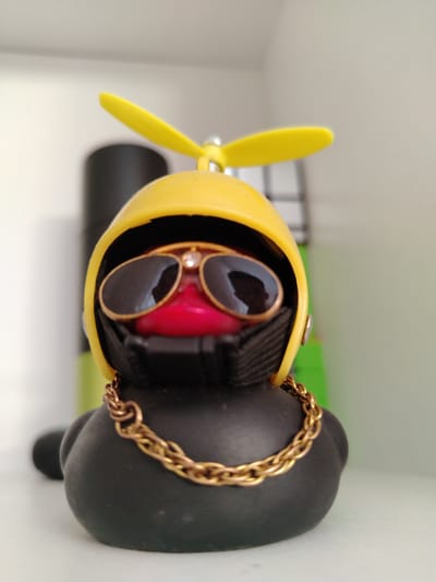 A close-up, shallow depth-of-field shot showcases a black rubber duck figurine adorned with accessories. The duck wears a bright yellow helmet with a yellow propeller attached to the top. It sports dark, reflective sunglasses with gold rims and a small clear gem between the lenses. A bright red, stylized beak is visible beneath the sunglasses. Around the duck's neck is a thick, braided gold chain. The figurine is positioned in the foreground, with a blurred background that suggests an indoor environment, possibly a shelf or display. The overall impression is that of a novelty item. This item is located in Slonim, Belarus.