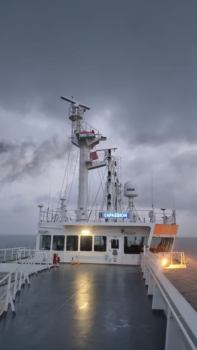 The image shows the upper deck of a vessel at sea, under a cloudy sky. A tall mast structure dominates the center, adorned with radar equipment and an Indian flag. The vessel's name, "SEAPASSION," is illuminated in blue lettering on its superstructure. The deck itself is wet, reflecting the overhead lights. In the background, dark smoke billows from the vessel's stack. The location is not specified as Pirotan, India, but the presence of the Indian flag suggests a possible connection to India.