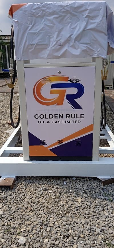 A new or recently installed white fuel dispenser, mounted on a white metal frame, is centrally positioned on a bed of light grey and tan gravel in Isheri-North, Nigeria. The top section of the dispenser is covered with a wrinkled white protective sheet, partially revealing an orange material underneath on the upper left. The dispenser's front panel displays the "GOLDEN RULE OIL & GAS LIMITED" logo, featuring a stylized orange "G" and blue "R," set against a white background with lower geometric patterns in orange and dark blue. A silver lock is visible on the panel. Black fuel hoses with metallic fittings, including a blue valve on the left hose and pressure markings "PETROTEC 10BAR / 150PSI," extend from both sides of the unit. A small wooden block rests on the gravel near the dispenser's base on the left. The background shows blurred green foliage and indistinct structures under a light sky. No individuals are present. The photograph was taken on January 10, 2026, at 13:36.