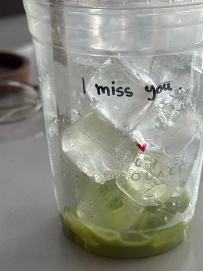 A plastic cup holds ice, a green drink, and is sealed with a light green lid. The cup has condensation and the phrase "I miss you." written on it in black marker. A small, red item sits on one of the ice cubes. The word "SOLACE" is visible through the liquid on the side of the cup. Another blurred object appears in the lower left corner of the image.