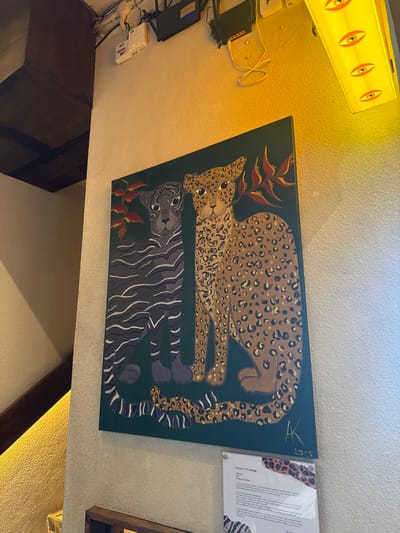 The image captures an interior scene featuring a prominent painting on a light-colored, textured wall. The painting is rectangular, oriented horizontally, and depicts two large feline creatures against a dark green background.
The upper animal is a leopard or jaguar, painted in shades of tan and brown with numerous black rosettes (spots). It is depicted lying down, facing left, with its head slightly raised and looking forward. Its eyes are dark, and its mouth is closed. Its tail curls towards the right side of the canvas.
Below the leopard, and mirroring its pose, is a tiger. This animal is rendered in dark grey with distinct black stripes. It is also lying down, but faces right, with its head raised and looking forward. Its eyes are dark, and its tail curls towards the left side of the canvas.
Stylized plant elements, possibly tropical flowers or leaves in shades of reddish-brown, are interspersed in the dark green background. One cluster appears near the leopard's head on the upper left, and another near the tiger's tail on the lower right. In the top right corner of the painting, a signature "AK" and the date "2023" are subtly visible.
The wall itself is a light, creamy off-white or beige color with a textured finish.
Above and to the left of the painting, a long, rectangular overhead light fixture emits a warm yellow glow. This fixture has several circular or oval cutouts that resemble eyes.
Further to the left, below the light fixture, several electronic devices, including black boxes and coiled white cables, are mounted on the wall. A white power strip is also visible.
To the right of the painting, an information sign is displayed in a transparent acrylic frame. The paper within the frame features abstract animal print patterns (leopard spots and zebra stripes) along its edges. The title "Natures of Change" is clearly readable at the top of the sign. Below the title, a paragraph of text provides context for the artwork, explaining that the leopard and tiger represent "strength, adaptability, and the interconnectedness of all living things," with the leopard's spots symbolizing "individuality and unique experiences," and the tiger's stripes signifying "unity and collective action."
In the lower part of the image, a dark brown, possibly wooden, rectangular object (like a piece of furniture or a shelf) is visible, along with a darker brown wooden beam or trim running diagonally.
The overall lighting in the scene is warm and bright, originating predominantly from the yellow light fixture, creating a cozy atmosphere.