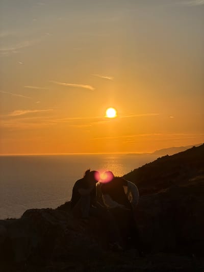 A silhouette of two individuals is captured in the foreground, sitting on a rocky outcrop overlooking the sea during sunset. The sun, a bright orb, is positioned in the center of the frame, casting a warm orange glow across the sky and water. The individuals are positioned close together, appearing to be sharing a moment. One individual, on the left, has their head bowed, with their hair tied up in a bun. The other individual, on the right, is leaning in towards the first. The sea stretches out to the horizon, reflecting the vibrant colors of the sunset. Hills with dark outlines are visible in the distance on the right side of the image. The overall scene depicts a tranquil and romantic natural setting.