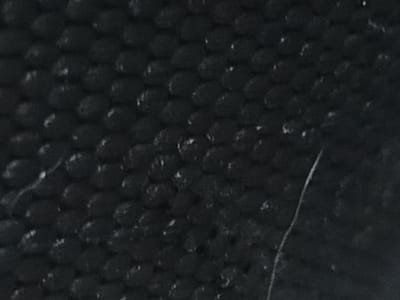 The image is a close-up, abstract shot of a textured black surface. It appears to be a coarse material with a pattern of raised, rounded bumps arranged in a hexagonal grid. Some of these bumps have small, light-colored speckles or imperfections on their surface, suggesting wear or dirt. There is also a thin, light-colored scratch or mark running diagonally across the lower right portion of the image, which stands out against the dark background. The overall impression is one of a weathered, tactile surface. No elements are visible to place this image within Al Hadaba, Egypt.