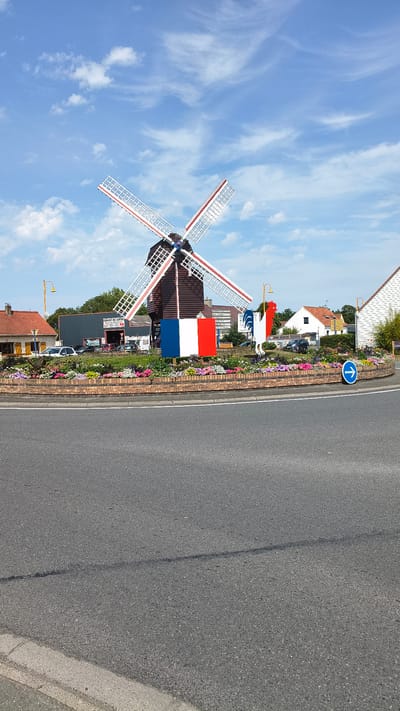 This image shows a roundabout in Marck, France, featuring a decorative windmill and a rooster statue. The windmill has brown wooden construction with white and red striped sails. A French flag is placed prominently at the base of the windmill. To the right of the windmill, a rooster statue, also painted in the colors of the French flag (blue, white, and red), stands. The roundabout is surrounded by a brick border filled with colorful flowers. Beyond the roundabout, buildings and trees are visible, including a building with a sign reading "GARAGE DU LOIRE". The sky is blue with scattered white clouds. A blue directional arrow sign indicating traffic flow is also present on the roundabout. The foreground shows a paved road.
