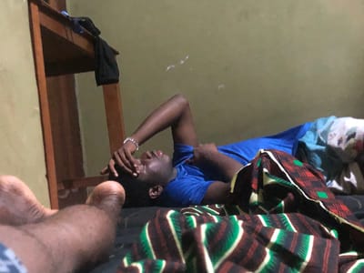 In Nwofia, Nigeria, a man is lying on a bed with a patterned blanket partially covering him. He is wearing a blue t-shirt and has his hand on his forehead. A wooden shelf stands beside the bed. Another person's feet are visible in the foreground. The background features a plain wall.