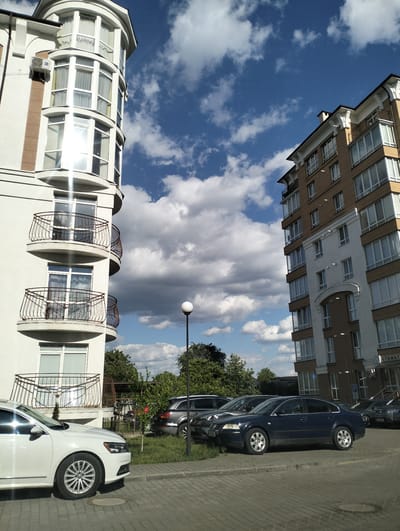 The image shows a street scene in Krykhivtsi, Ukraine. Two multi-story buildings frame a view of a partly cloudy sky. The building on the left is white with a rounded corner featuring several balconies. The building on the right is a mix of tan and white with a more traditional rectangular design. A parking lot is visible in front of the buildings, occupied by several cars, including a white sedan on the left and a dark blue sedan towards the center. There is a lamppost in front of trees between the two buildings. The ground is paved with gray paving stones. The signage near the right building indicates a possible стоматолог (dentist) location.
