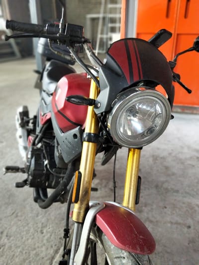 A close-up, low-angle, head-on shot features the front of a red and black motorcycle parked indoors. The motorcycle has gold-colored front suspension forks and a round, chrome-rimmed headlight. A small, black windscreen with red stripes is positioned above the headlight. The motorcycle's handlebars are visible on the left, with black grips and levers. The fuel tank is a prominent red color with black accents and appears to be a matte finish. Parts of the engine and frame are visible below the tank. The motorcycle is situated on a concrete floor, and the background includes out-of-focus orange industrial doors and shelving units, suggesting a workshop or storage area. There are no people visible in the frame. The lighting is natural and appears to be coming from the right side of the frame, casting shadows on the left. The image is not specific to Semey, Kazakhstan, as there are no identifying markers of this location.
