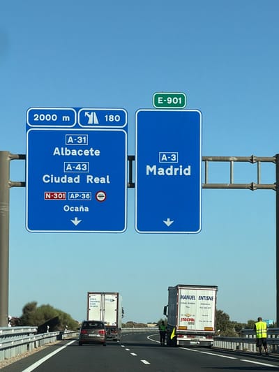 The image shows a highway directional sign in Tébar, Spain. The sign indicates that A-31 for Albacete is 2000 meters away and has an exit of 180 km. It also shows A-43 for Ciudad Real and N-301/AP-36 for Ocaña. To the right, another sign indicates A-3 for Madrid with a downward arrow. The sky is clear blue. In the background, there are vehicles on the highway, including trucks and cars, and vegetation along the roadside. Two individuals in high-visibility vests are present near the trucks.