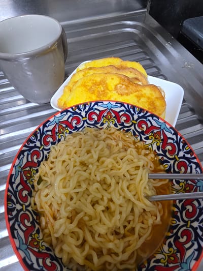 A top-down shot shows a meal in a kitchen setting. The primary subjects are a bowl of ramen noodles with chopsticks, a plate of french toast, and a gray mug. The ramen is in a colorful patterned bowl with red, blue, and yellow designs. The french toast appears to be golden brown and stacked on a white square plate. The gray mug is partially visible on the left side of the image, tilted as if it had been moved. The meal is placed on a stainless steel sink with a ribbed texture, a common sight in a kitchen. The lighting suggests it could be daytime, as there are no artificial lights clearly visible. The overall scene conveys a sense of a home-cooked meal, possibly breakfast or lunch, in South Korea, given the location context. There is no text visible in the image.