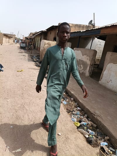 The scene depicts a young male walking on an unpaved dirt road in Zango, Nigeria. The subject, a Black male appearing to be in his late teens or early twenties, walks directly towards the camera, maintaining eye contact. He wears a knee-length, long-sleeved, loose-fitting green tunic with a buttoned V-neck and a breast pocket, over matching green trousers and red and brown flip-flops. His shadow is cast to his left on the dirt ground. To the subject's right, an open drainage ditch or gutter is filled with assorted refuse, including plastic bags and wrappers. In the background, a series of single-story, utilitarian buildings line both sides of the road, constructed from mud brick or concrete, many with corrugated metal roofs. A grey horn-shaped loudspeaker is visible on a pole atop one of the buildings to the right. Distant figures and a cart are discernible further down the road. The sky is clear and bright.