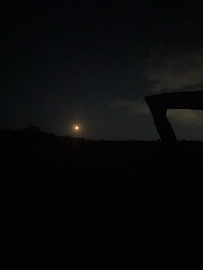 A dimly lit night scene captured from a low angle depicts a dark silhouette of a hilly landscape against a night sky. A bright, yellowish moon is visible near the center-left of the frame, positioned above the dark horizon. To the right, a dark, angular object, possibly a fence or a structure, is silhouetted against the sky, with a portion of cloudy sky visible behind it. The foreground is entirely black, suggesting it is either too dark to capture detail or a dark surface. The overall impression is one of a serene, possibly rural, night. The image is devoid of any visible people, actions, or interactions.